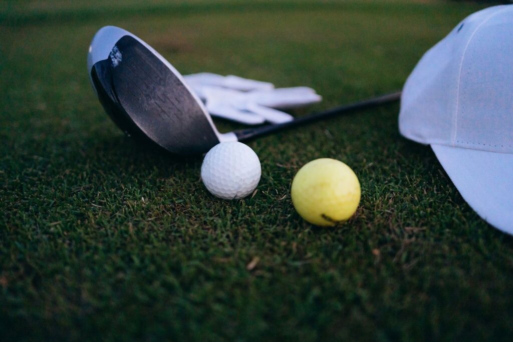 Golf balls and clubs resting on closely cut grass beside a white cap