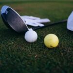Golf balls and clubs resting on closely cut grass beside a white cap