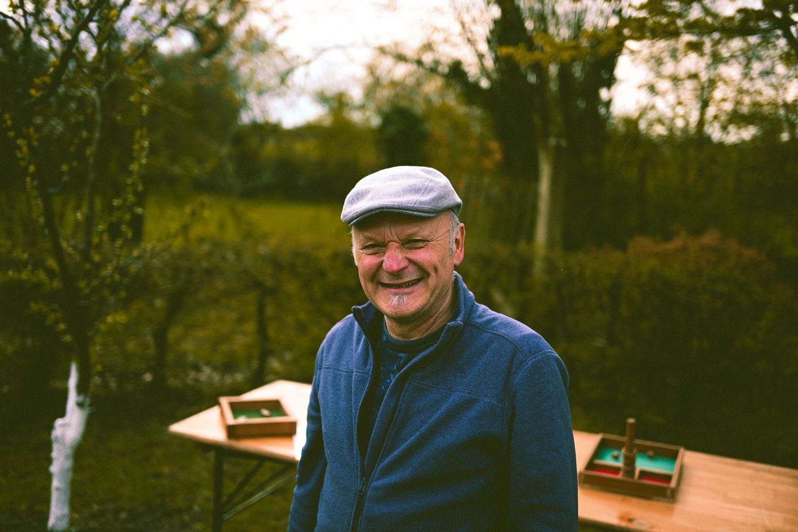 Smiling older man in a flat cap standing beside outdoor game tables